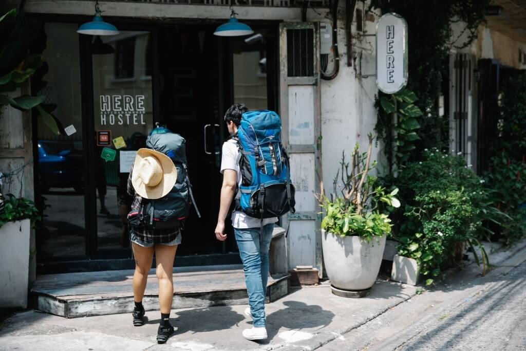 Casal de mochileiros caminha para a entrada de um hostel. Imagem disponível em Pexels.