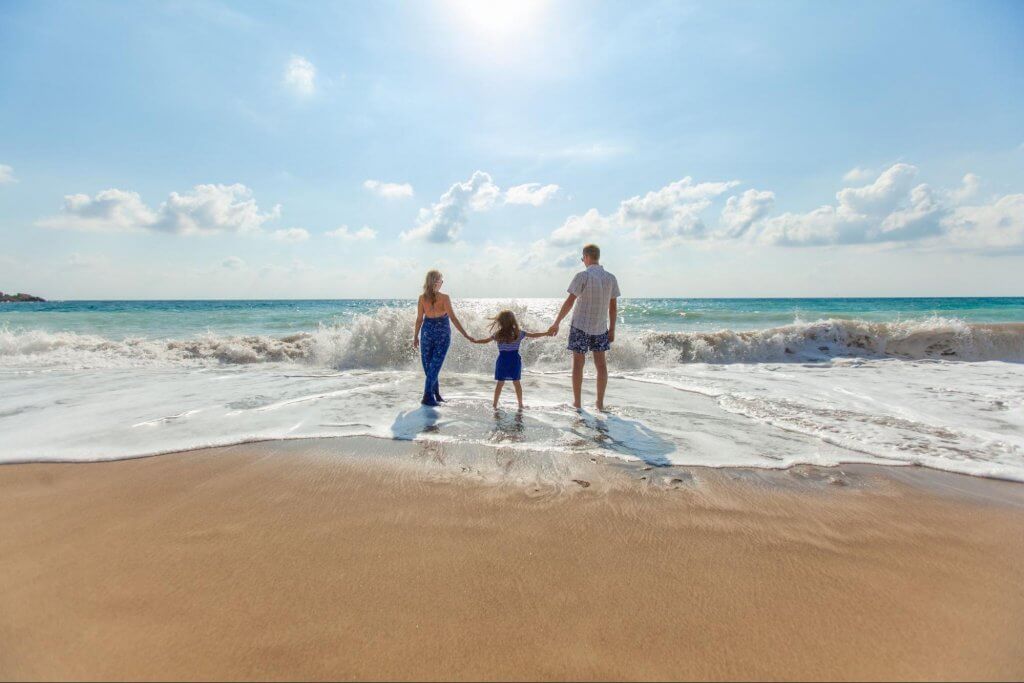 imagem de uma família, na beira da praia, de mãos dadas. Homem, mulher e uma criança veem as ondas