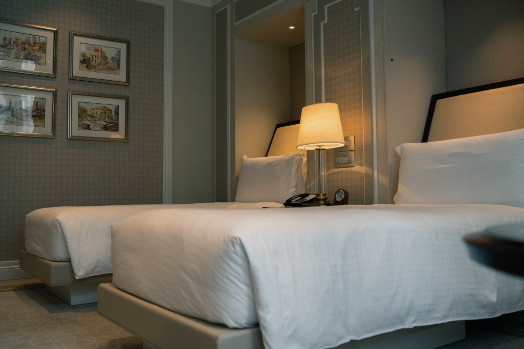 Cama de hotel de casal, com roupa de cama branca e abajur ao fundo. Imagem disponível no Unsplash