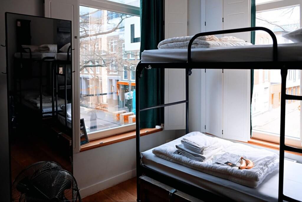 quarto de hotel em estilo dormitório. Imagem mostra uma beliche, com roupa de cama e itens pessoais. Em segundo plano, uma janela mostrando a cidade