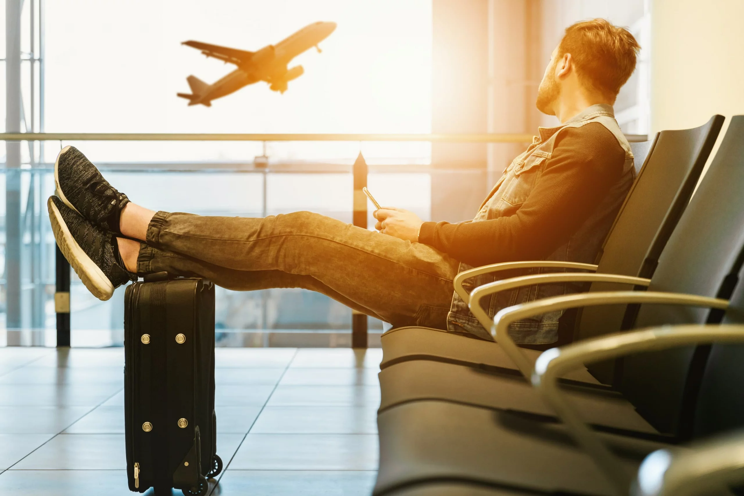Homem sentado no saguão de um aeroporto vê um avião decolar. Imagem disponível em Unsplash.