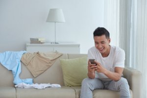 homem de camiseta branca sentado em uma sofá sorri enquanto usa seu celular