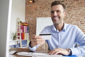 homem sentado ao computador sorri enquanto segura um cartão de crédito
