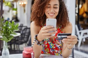 mulher sentada a uma mesa sorri enquanto segura um cartão de crédito e um celular