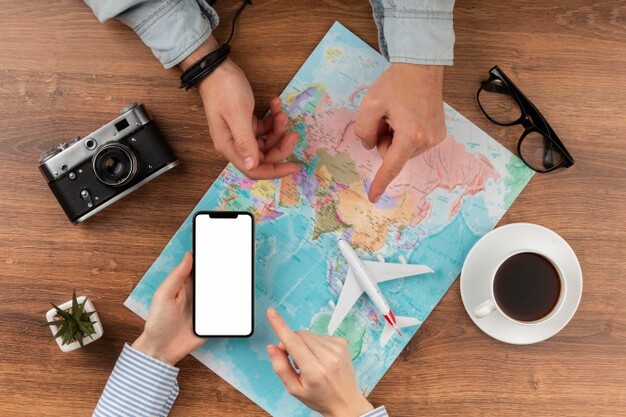 imagem ampliada e vista de cima das mãos de duas pessoas em lados opostos de uma mesa de madeira com um mapa, um avião em miniatura, um par de óculos, uma câmera, uma xícara de café e um pequeno vaso com um cacto