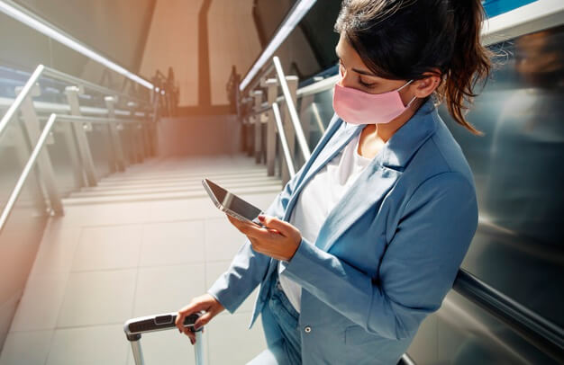 mulher de blazer azul e máscara mexe em seu celular enquanto segura sua bagagem no topo de uma escadaria