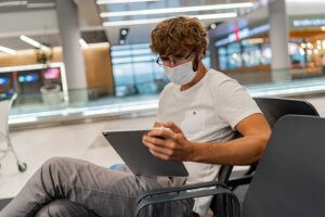 homem de máscara usando um tablet sentado em uma cadeira de aeroporto