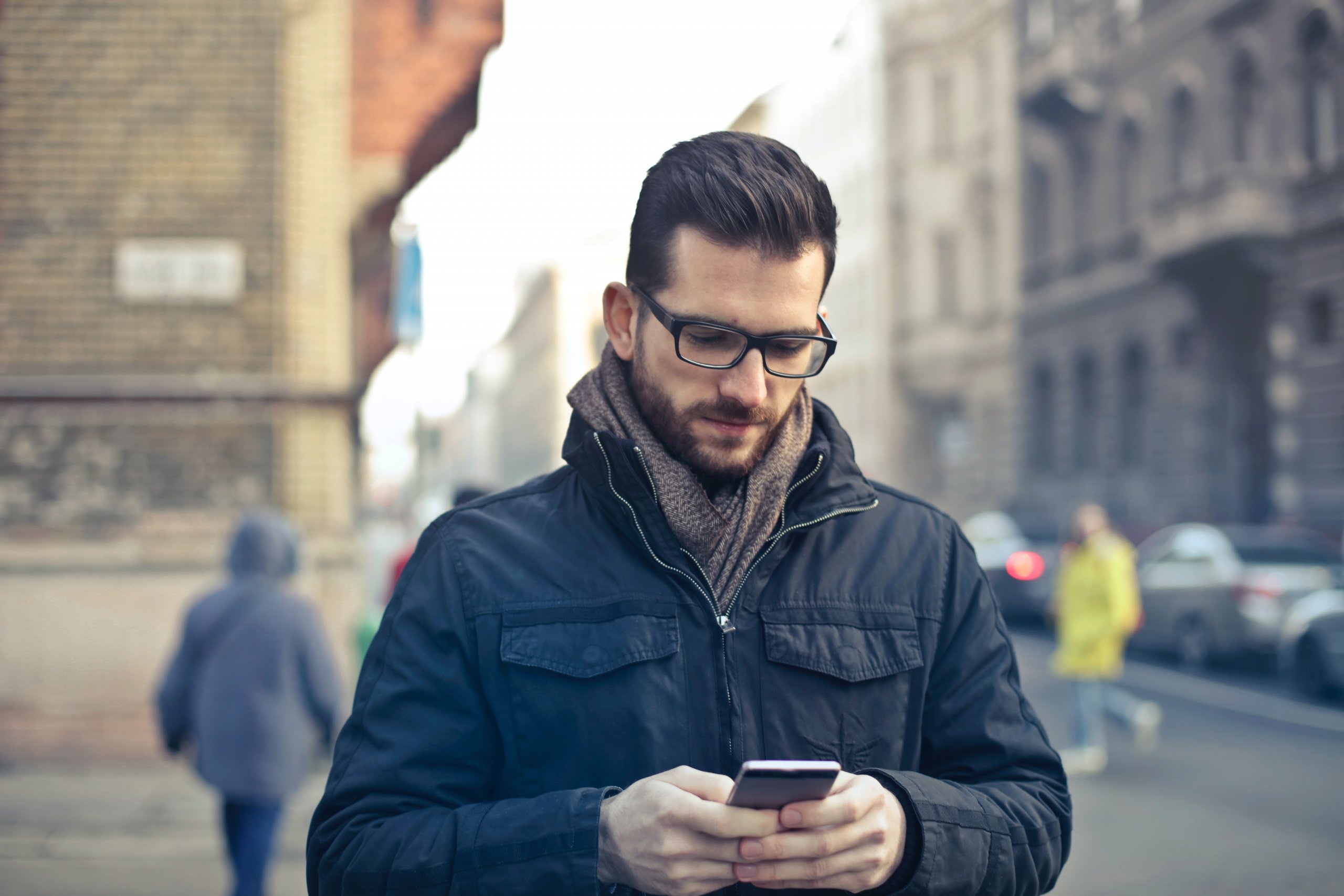 Homem Vestindo Uma Jaqueta Preta Com Zíper Segurando Um Smartphone Cercado Por Prédios De Concreto Cinza. Disponível em Pexels