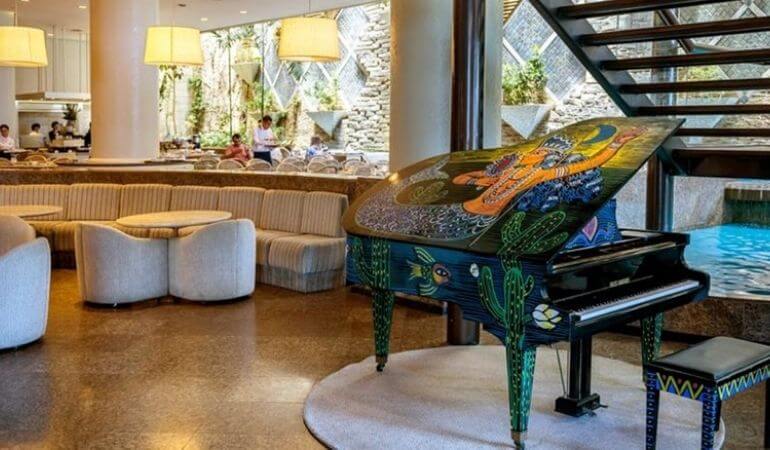 um piano decorado, próximo a poltronas e mesinhas e, aos fundos, a área de restaurante do hotel