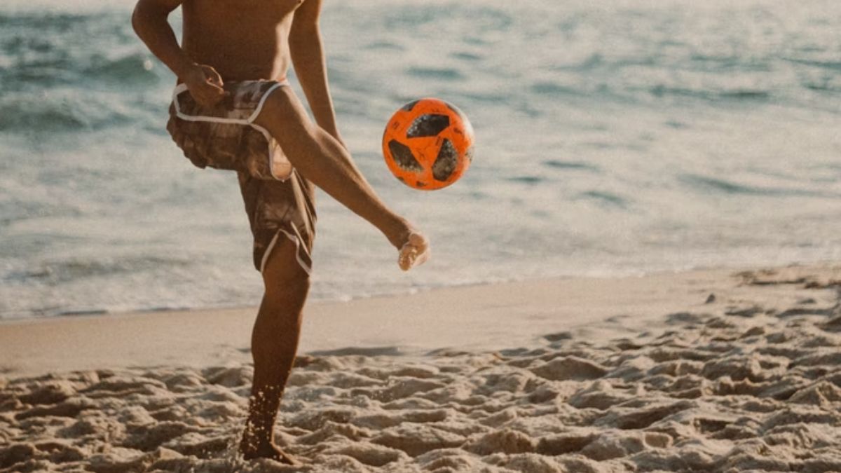 pessoa jogando uma bola de futebol na areia beira da praia