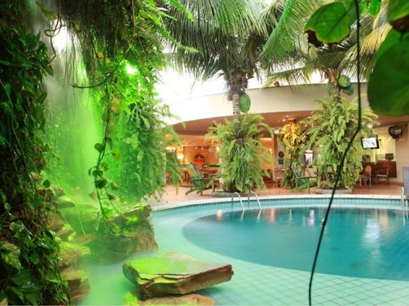 Vista da cascata e da piscina do Amazon Plaza hotel, com cadeiras e área de descanso ao fundo, e vegetação à frente na foto