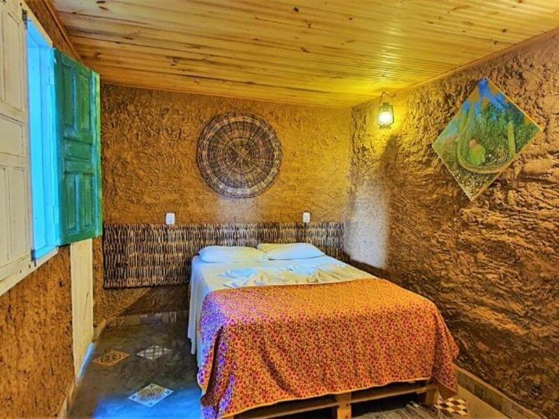 Quarto do Vila Aju, com cama de casal com roupa de cama em tom laranja. As paredes e teto tem tons e texturas terrosos