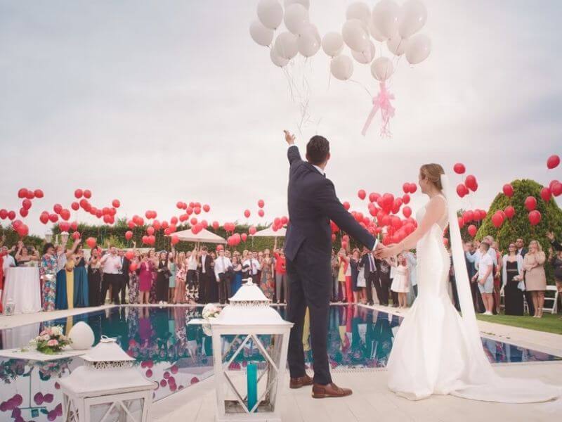 marido e mulher jogando balões em meio a um destination wedding em local com piscina e várias pessoas ao redor.