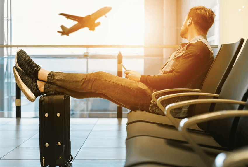 Homem sentado no saguão de um aeroporto, com os pés em cima da mala, observando a decolagem de aviões