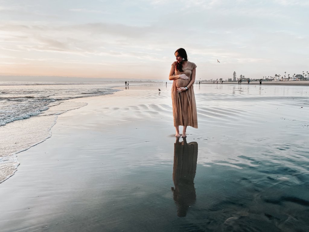Mulher grávida, usando um vestido marrom, fica em pé próximo ao mar. Imagem disponível em Unsplash.