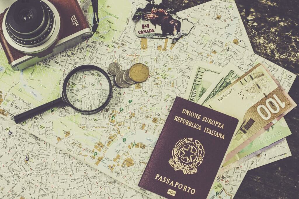 Passaporte em cima de um mapa, ao lado de lupa, câmera e moedas. Imagem disponível em Unsplash.